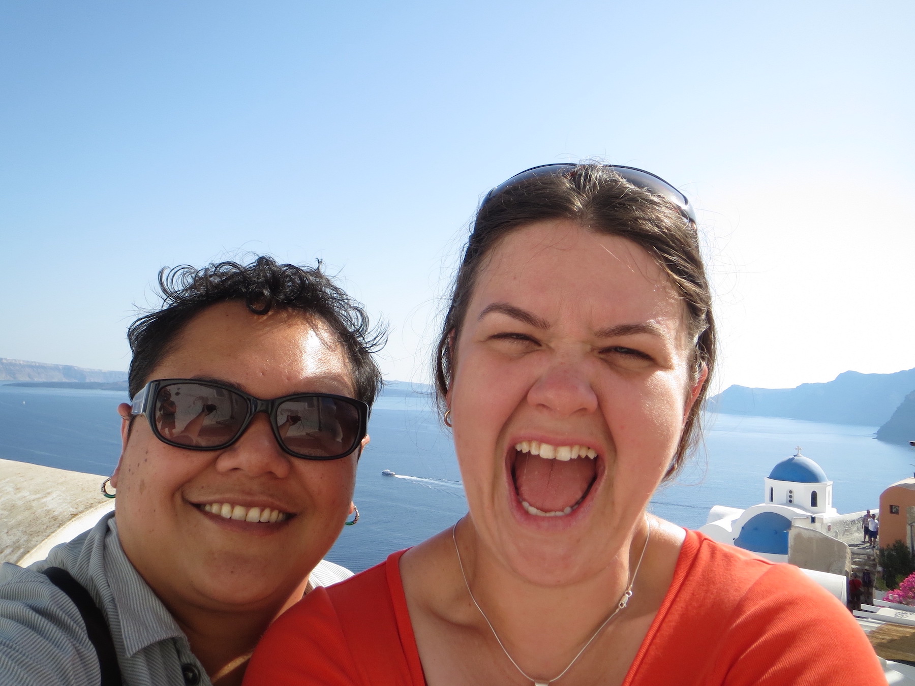 Maylene & Charity soaking the sun at the famous scenic outlook in Oia, Santorini, Greece. Whitewashed homes and blue roofs cling to cliffs above the blue sea-filled crater below
